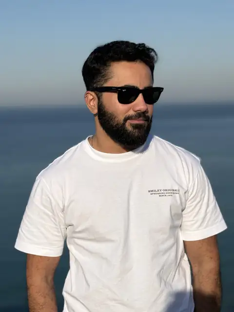 Emre Kent wearing sunglasses and a white T-shirt, photographed with the sea in the background.